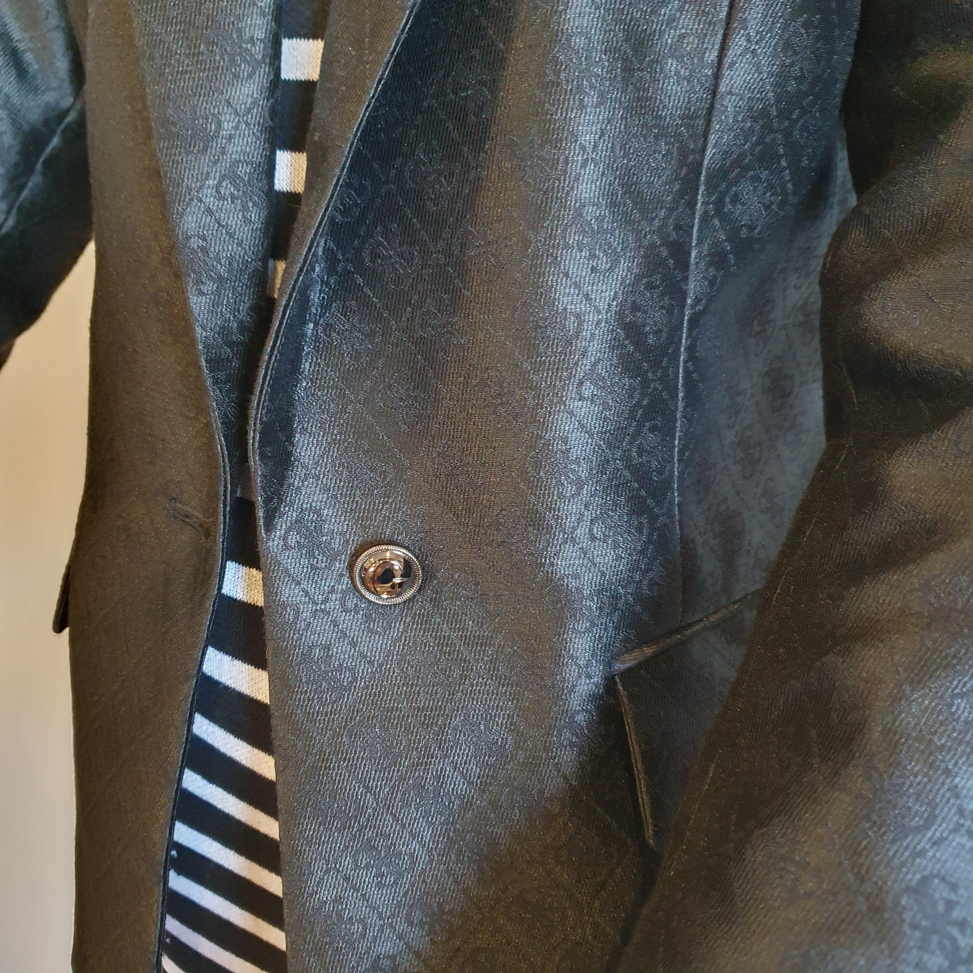 Blazer Guess monogrammé noir, détail du revers — rayures de la robe graphique entrevues en arrière-plan — Élégance Intemporelle