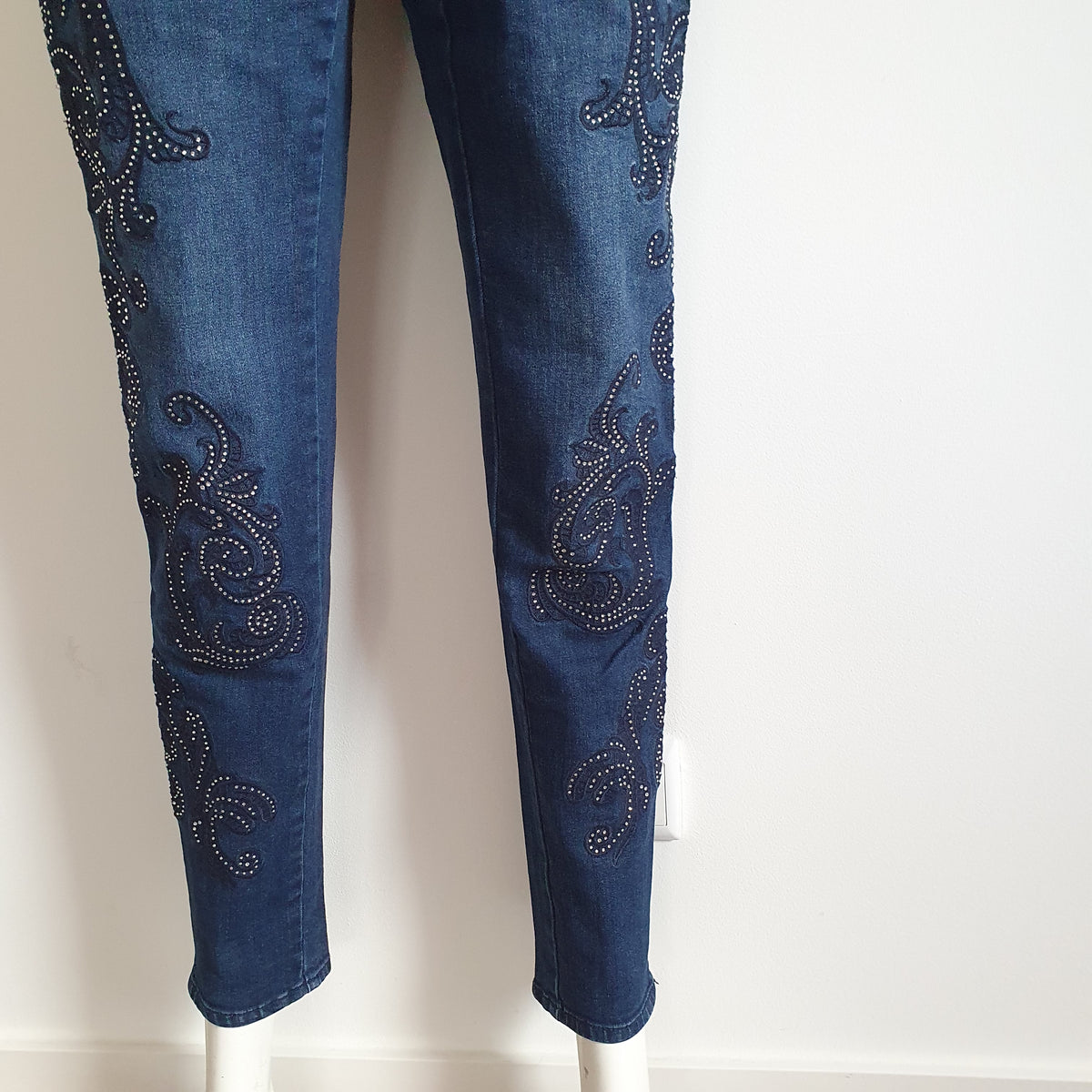 Gros plan des motifs arabesques à strass et des ourlets du jean bleu Madeleine sur les deux jambes. 