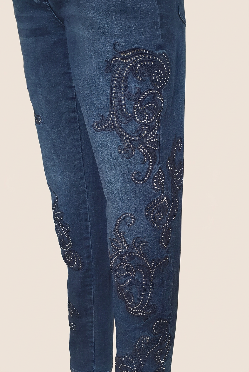 Détail des arabesques sombres et des strass sur la jambe du jean bleu Madeleine, vue de trois quarts.