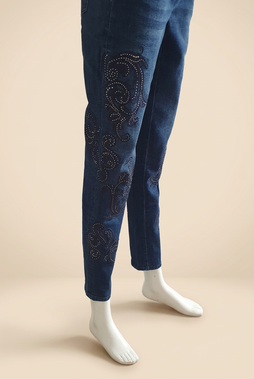Jean bleu Madeleine vu de trois quarts sur mannequin blanc, arabesques et strass le long des jambes, fond beige clair.